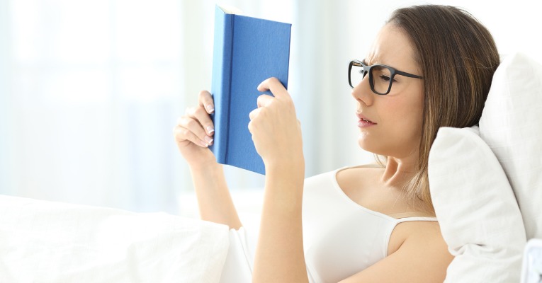 femme allongée dans le lit en train de lire un livre avec position de lecture avec trop oreiller et risque de douleurs dorsales type dorsalgie 