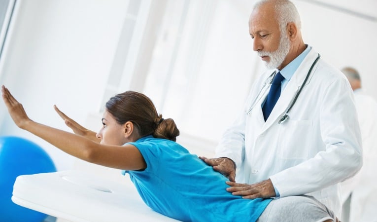docteur guide une femme allongée sur le ventre lors d'un exercice de rééducation renforcement du dos pour soigner lombalgie chronique