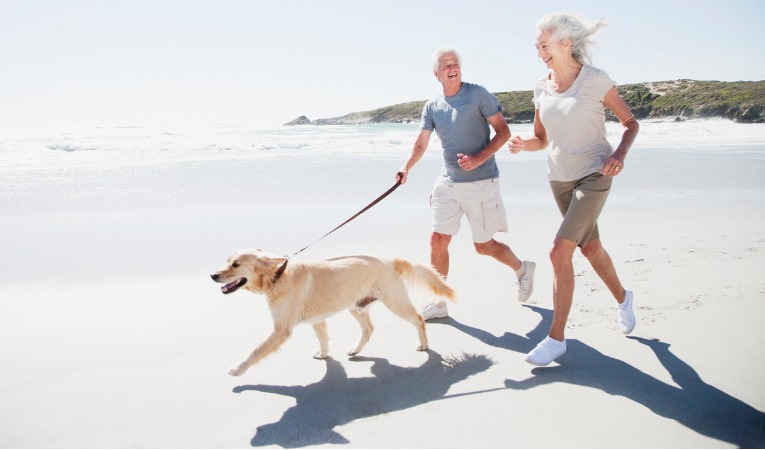 couple de sénior souriant qui court sur une plage avec leur chien après chirurgie de prothèse de genou
